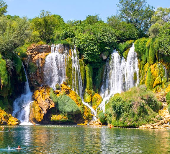 Kravica Waterfall in Bosnia with lush greenery and people swimming in the clear water.