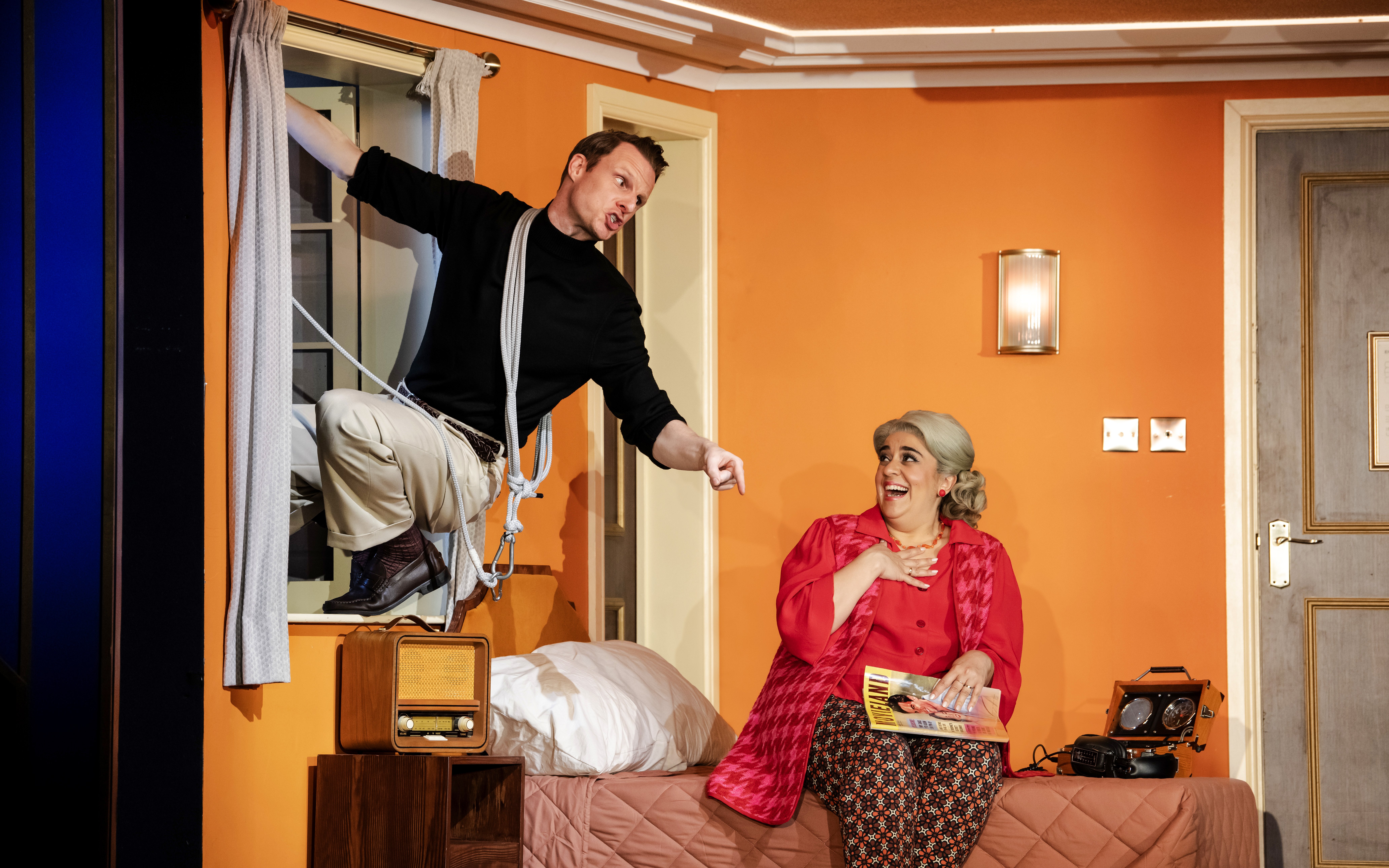 Man climbing through window with rope, woman sitting on bed laughing, scene from "The Comedy About Spies.
