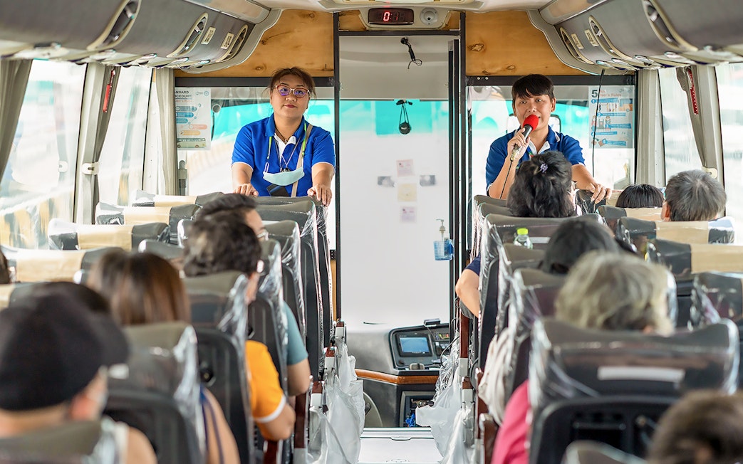 Tour guide speaking to passengers on a bus for Damnoen Saduak Floating Market tour.