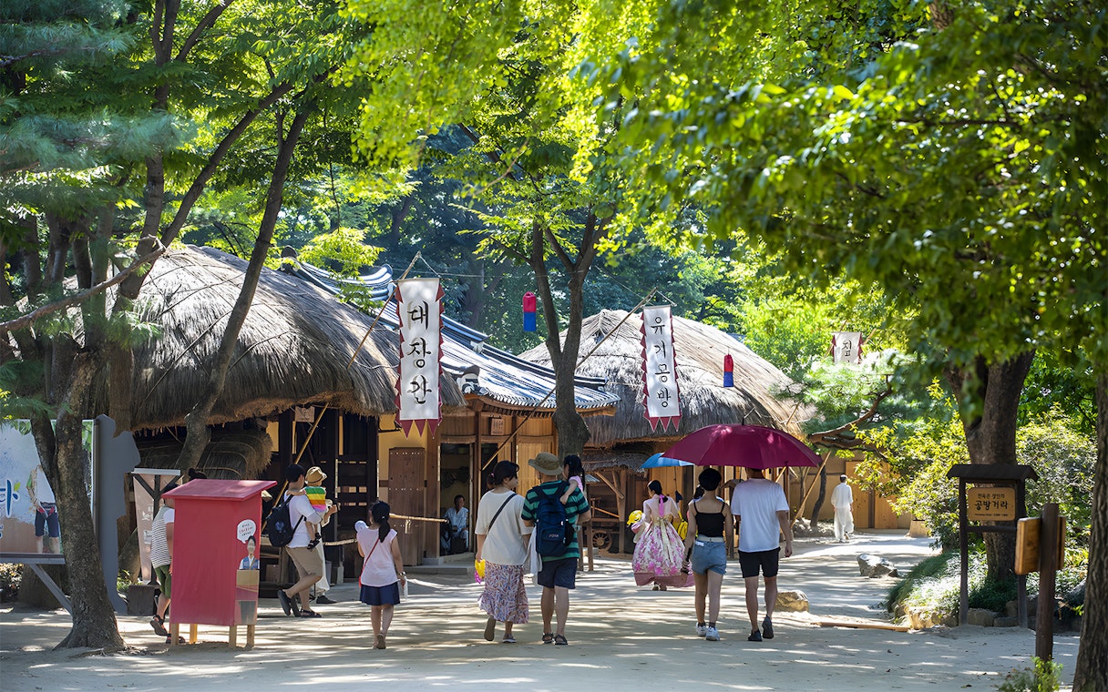 Visitors walking through traditional Korean Folk Village with thatched-roof houses, Suwon Hwaseong tour.