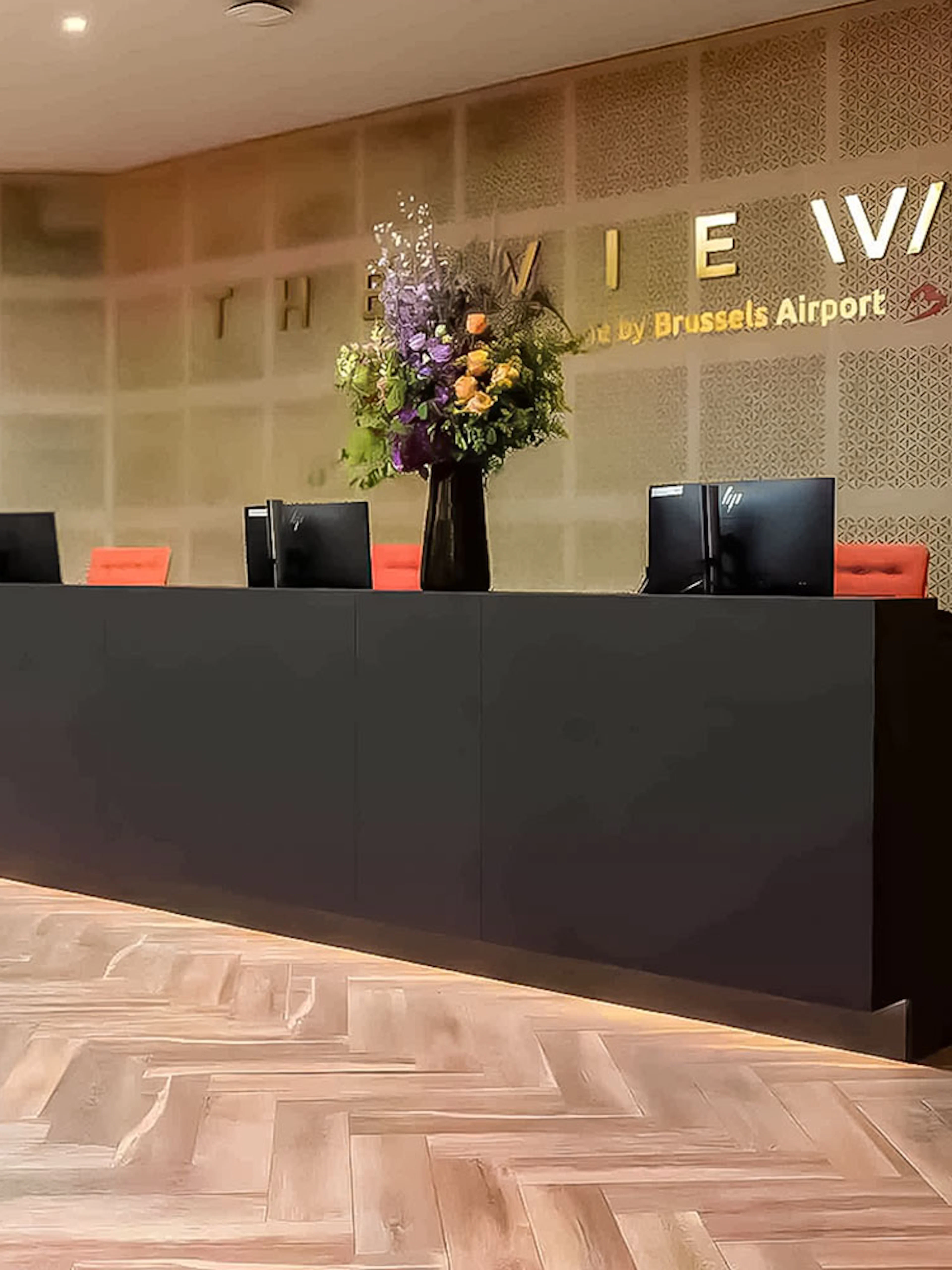 Reception desk at The View Brussels Airport Lounge with computers and floral arrangement.