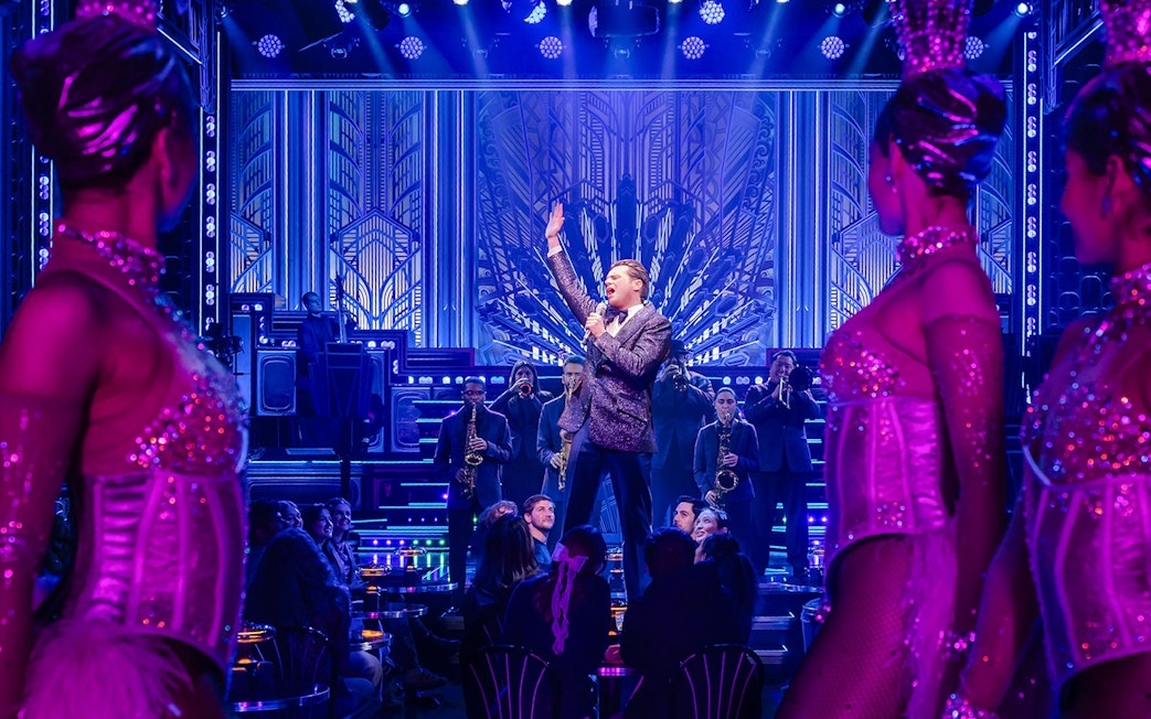 Performer on stage with band and dancers in vibrant theater setting.