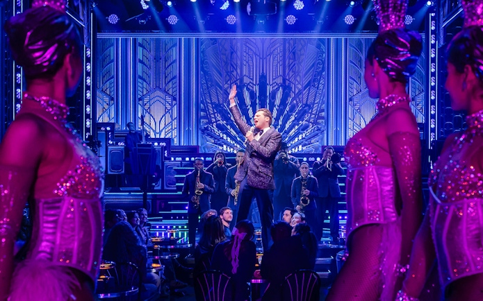 Performer on stage with band and dancers in vibrant theater setting.