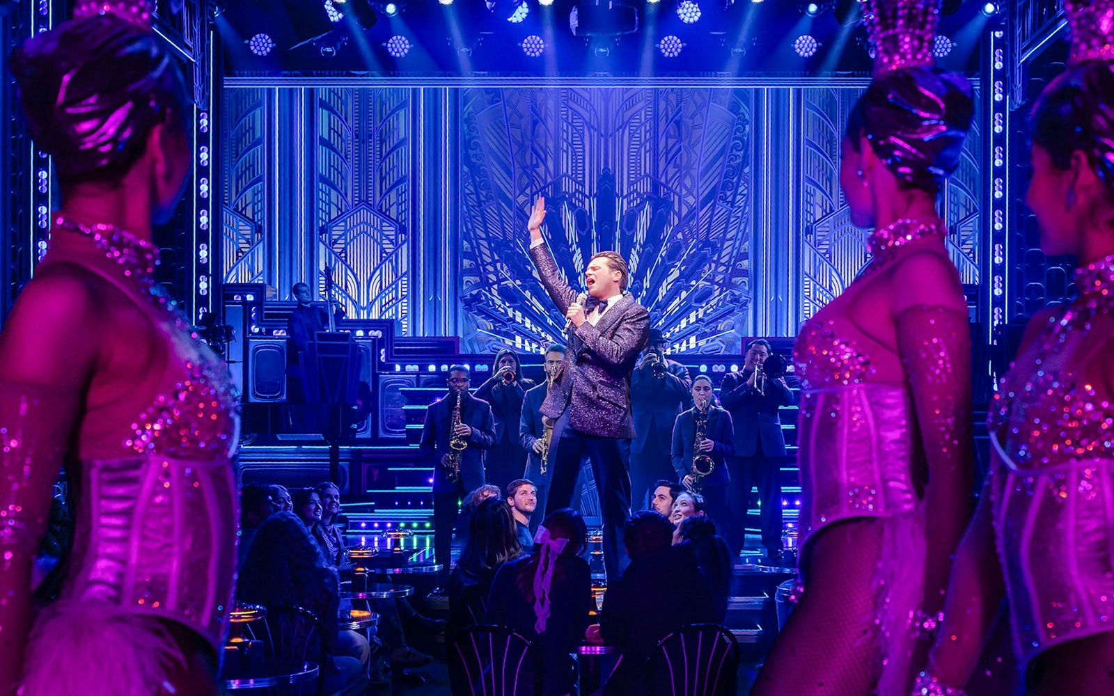Performer on stage with band and dancers in vibrant theater setting.