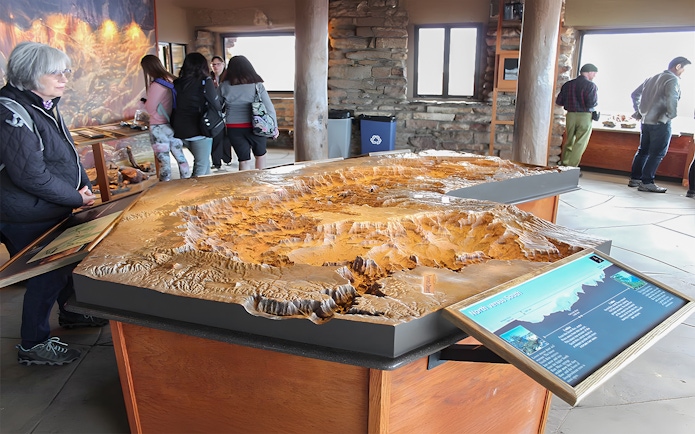 Grand Canyon South Rim visitors examining a detailed topographic model in an indoor exhibit.