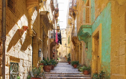 Narrow street in one of the Three Fortified Cities, lined with historic stone buildings and potted plants.