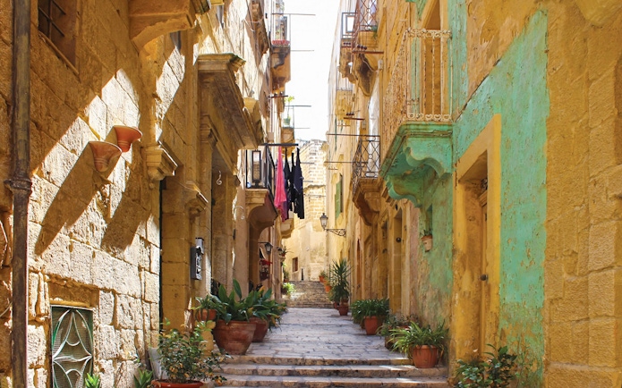 Narrow street in one of the Three Fortified Cities, lined with historic stone buildings and potted plants.