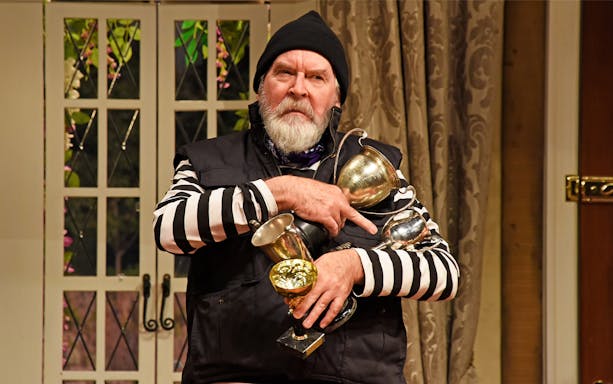 Man in striped shirt holding multiple trophies in a scene from "Noises Off" play.