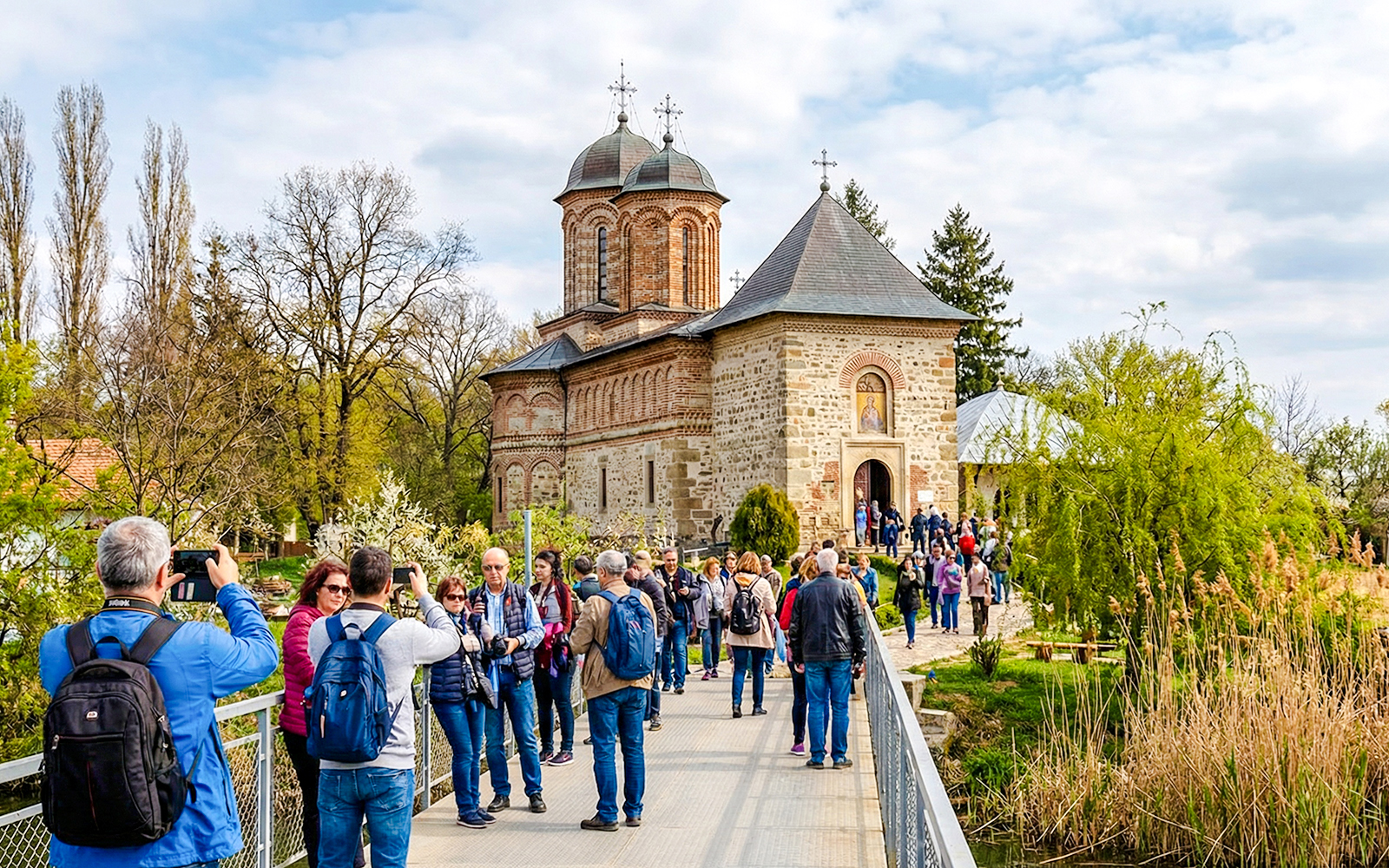 Guests entering Snagov Monastery, Romania, via a bridge surrounded by trees.
