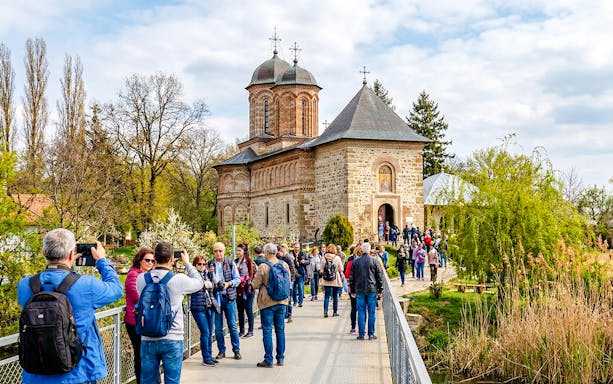 Guests entering Snagov Monastery, Romania, via a bridge surrounded by trees.
