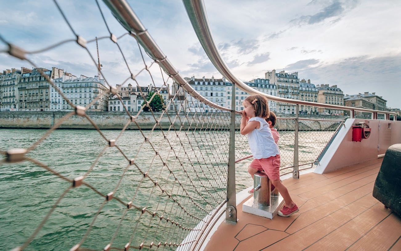 Children enjoying a Seine River cruise in Paris with historic buildings in the background.