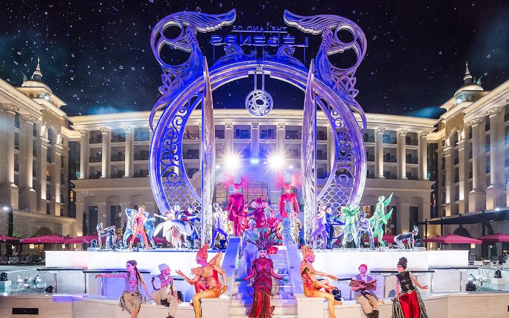 Performers in vibrant costumes at The Land of Legends boat parade, illuminated stage backdrop.