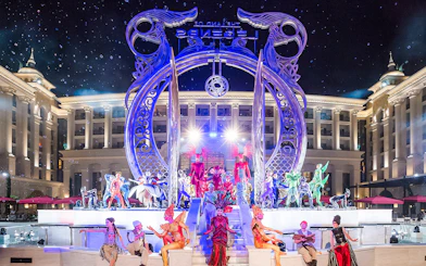 Performers in vibrant costumes at The Land of Legends boat parade, illuminated stage backdrop.