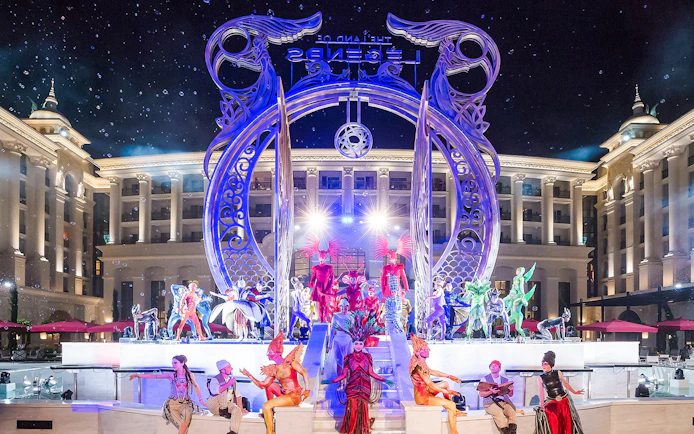 Performers in vibrant costumes at The Land of Legends boat parade, illuminated stage backdrop.