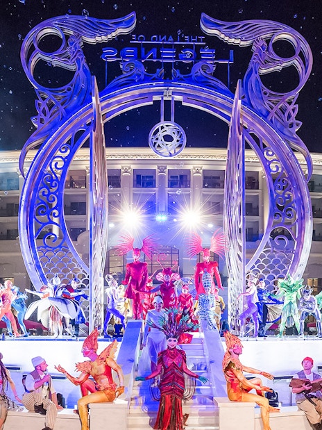 Performers in vibrant costumes at The Land of Legends boat parade, illuminated stage backdrop.