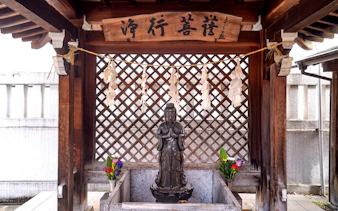Samurai statue at a Tokyo shrine with traditional wooden architecture and floral offerings.