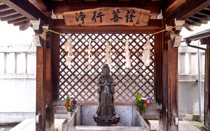 Samurai statue at a Tokyo shrine with traditional wooden architecture and floral offerings.