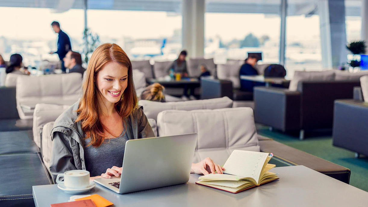 Woman using laptop in airport lounge with coffee and notebook nearby.