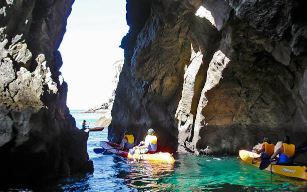 Kayakers paddling in clear waters of Arrábida Natural Park, Lisbon, with rocky cliffs in the background.