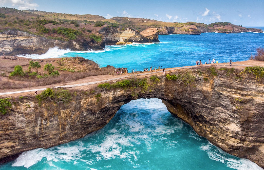 Tourists on a cliff at Broken Beach, West Nusa Penida, Indonesia, overlooking turquoise waters.