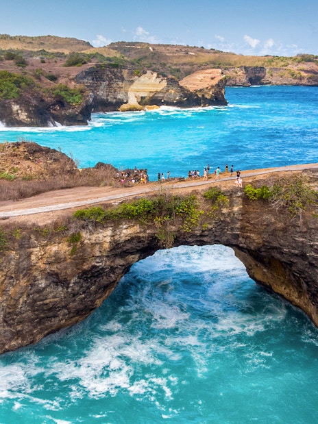 Tourists on a cliff at Broken Beach, West Nusa Penida, Indonesia, overlooking turquoise waters.