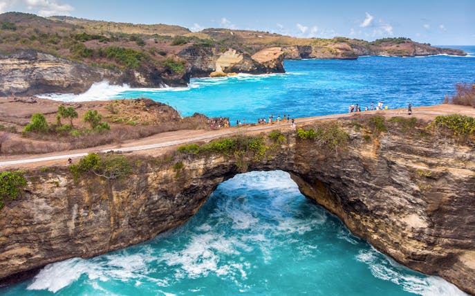 Tourists on a cliff at Broken Beach, West Nusa Penida, Indonesia, overlooking turquoise waters.