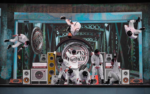 Dancers performing acrobatic stunts on stage at Global Village with speakers and urban backdrop.