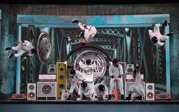 Dancers performing acrobatic stunts on stage at Global Village with speakers and urban backdrop.