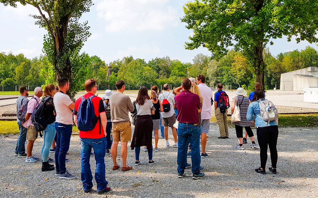 Visitors touring Dachau Concentration Camp grounds, surrounded by trees and historical structures.
