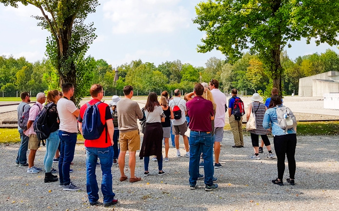 Visitors touring Dachau Concentration Camp grounds, surrounded by trees and historical structures.