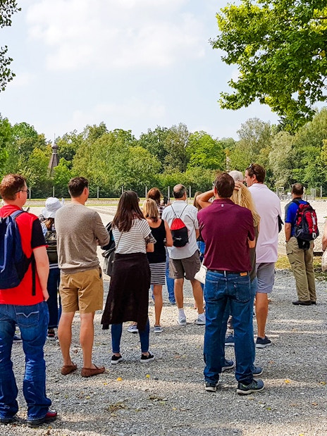 Visitors touring Dachau Concentration Camp grounds, surrounded by trees and historical structures.