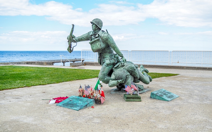 Statue of soldiers at Normandy D-Day Beach with flags and memorial plaques.