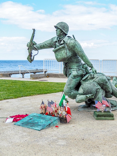 Statue of soldiers at Normandy D-Day Beach with flags and memorial plaques.