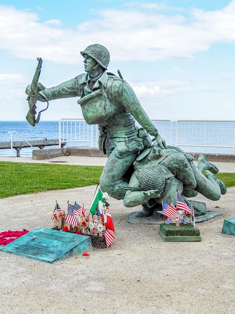 Statue of soldiers at Normandy D-Day Beach with flags and memorial plaques.