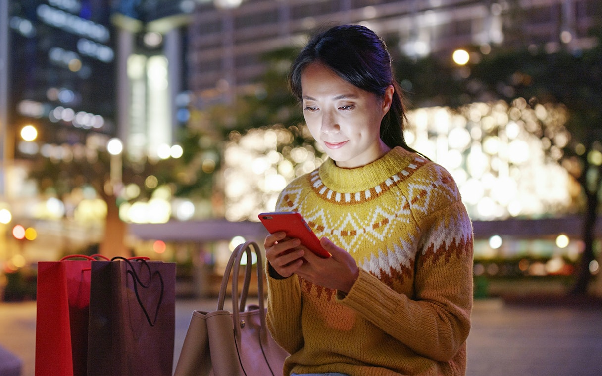 Woman using a smartphone in a city at night with shopping bags nearby.
