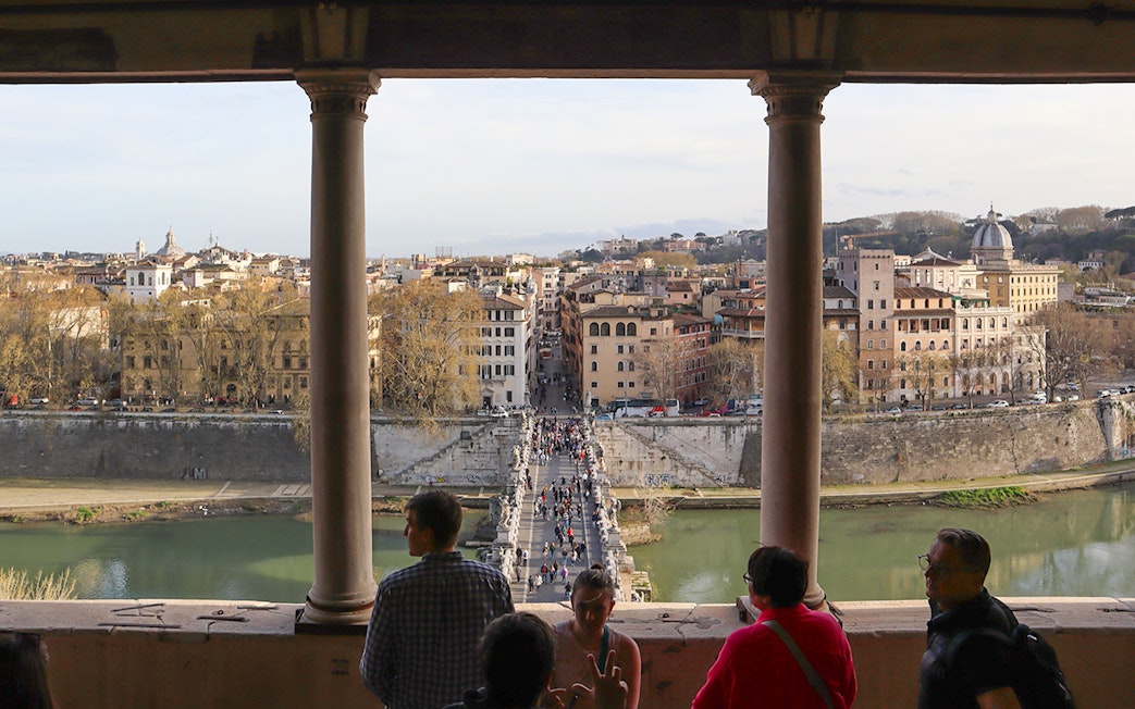 People walking on St. Angelo Bridge with a view of Rome's cityscape from Castel Sant'Angelo.