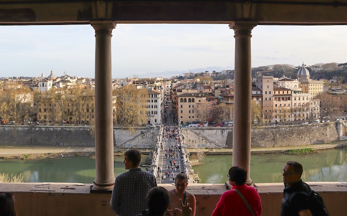 People walking on St. Angelo Bridge with a view of Rome's cityscape from Castel Sant'Angelo.