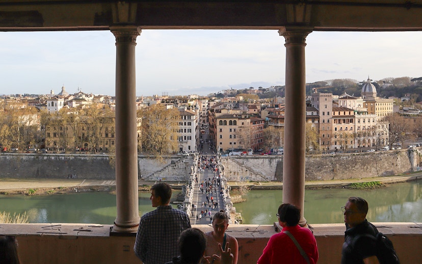 People walking on St. Angelo Bridge with a view of Rome's cityscape from Castel Sant'Angelo.