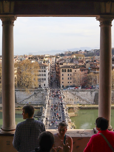 People walking on St. Angelo Bridge with a view of Rome's cityscape from Castel Sant'Angelo.