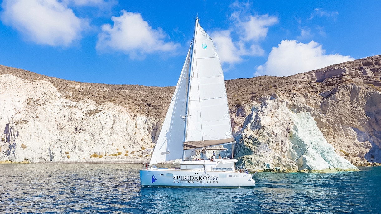 Catamaran sailing near rocky cliffs on Santorini luxury cruise.
