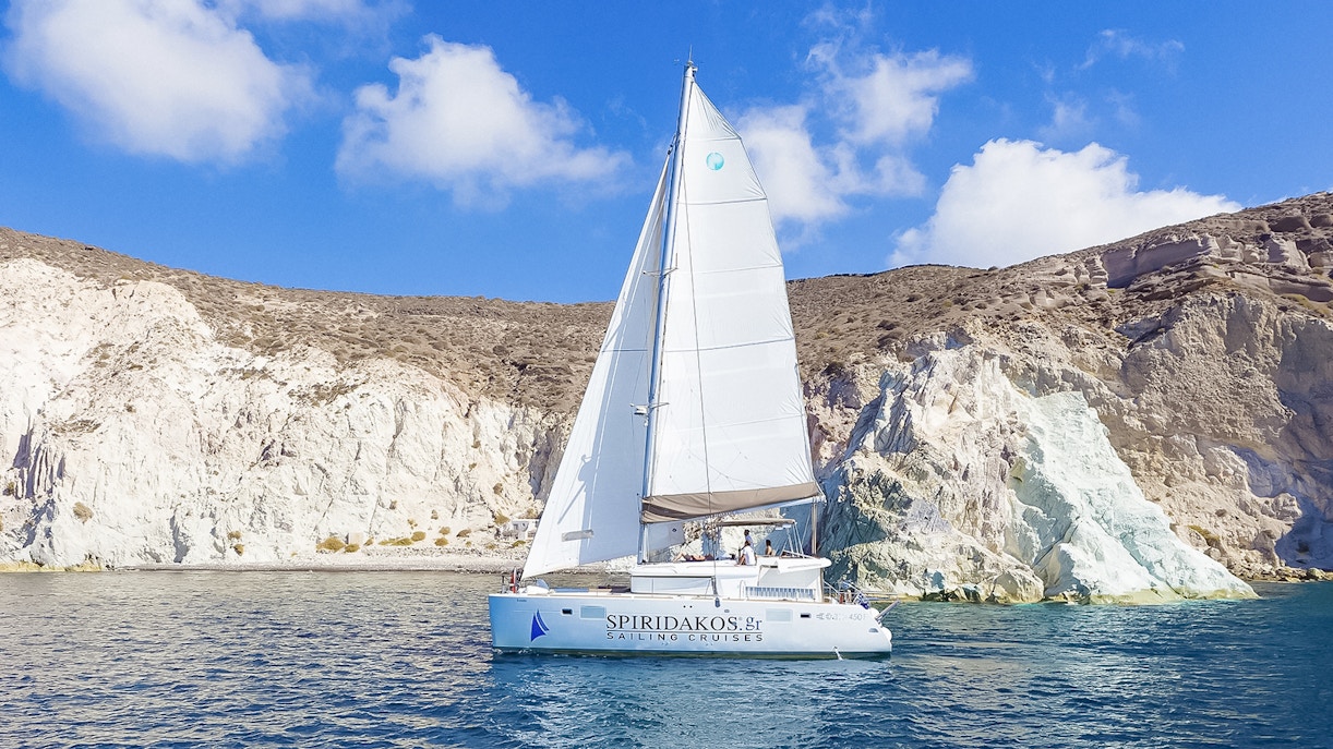 Catamaran sailing near rocky cliffs on Santorini luxury cruise.