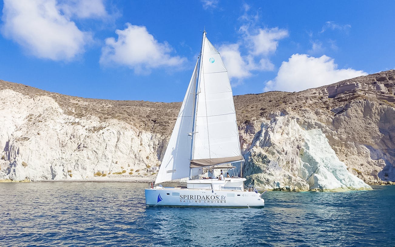 Catamaran sailing near rocky cliffs on Santorini luxury cruise.