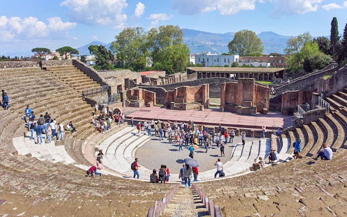 Tourists exploring the ancient amphitheater ruins in Pompeii under a clear blue sky.