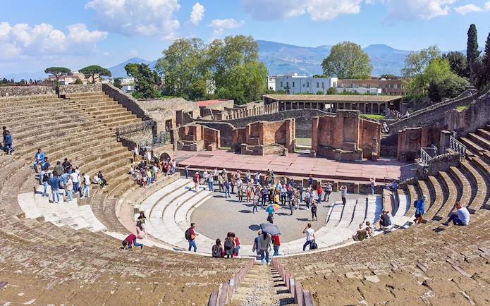 Tourists exploring the ancient amphitheater ruins in Pompeii under a clear blue sky.