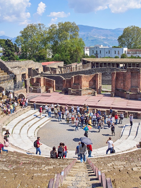 Tourists exploring the ancient amphitheater ruins in Pompeii under a clear blue sky.