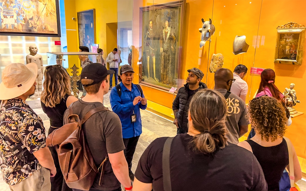 Tour group listening to a guide in the Anthropology Museum, Mexico, with artifacts and paintings displayed.