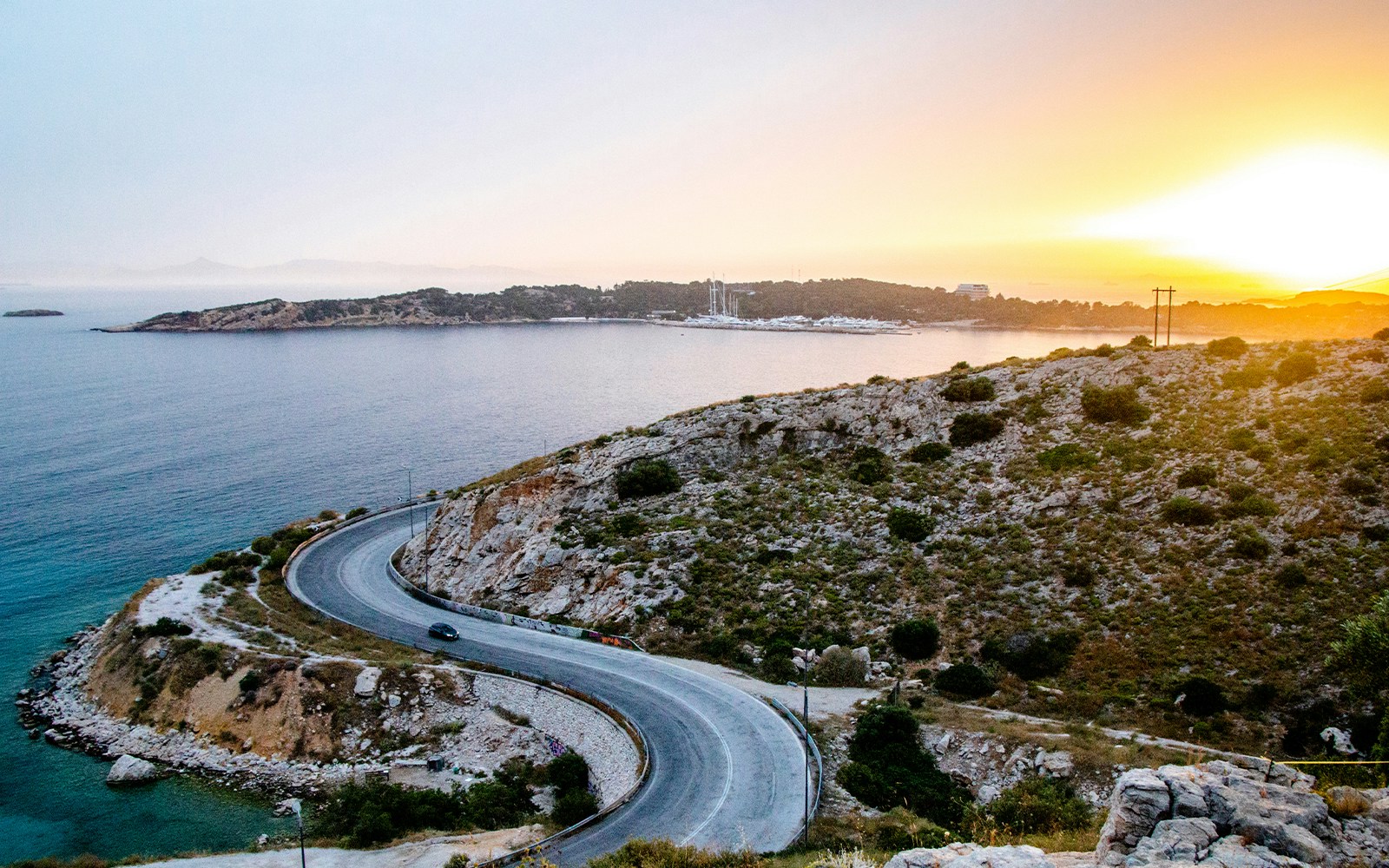 Serpentine road by the sea at sunset in Athens, Greece, with mountains and rocks.