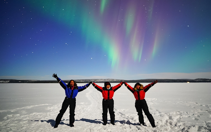 Tourists holding hands under Northern Lights in snowy landscape.