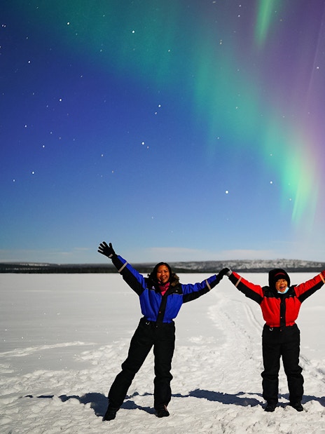 Tourists holding hands under Northern Lights in snowy landscape.