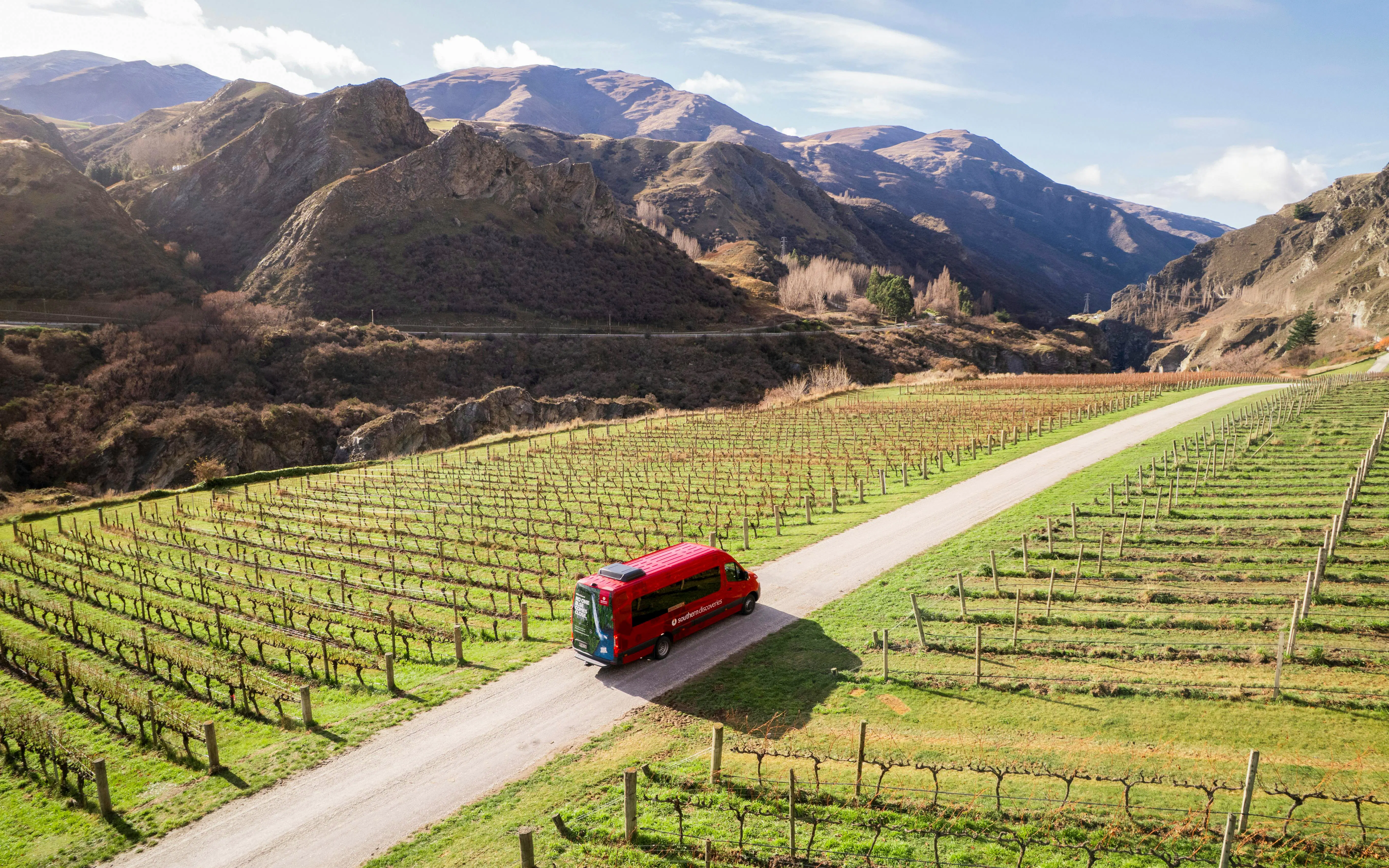 Red tour van driving through vineyard in Queenstown with mountain backdrop.